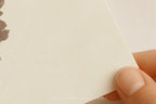 A close-up of a hand holding the corner of a textured off-white paper card or notebook page, with a small brown leaf illustration partially visible on the left side.