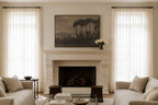 A styled living room featuring a large framed landscape painting above a white stone fireplace. The room is symmetrically arranged with sheer white curtains on both sides, light beige sofas, and a glass coffee table in the foreground.
