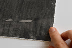 A close-up of a dark charcoal gray textured sheet of handmade paper with a rough, torn edge, held by a person's fingers against a clean white background.