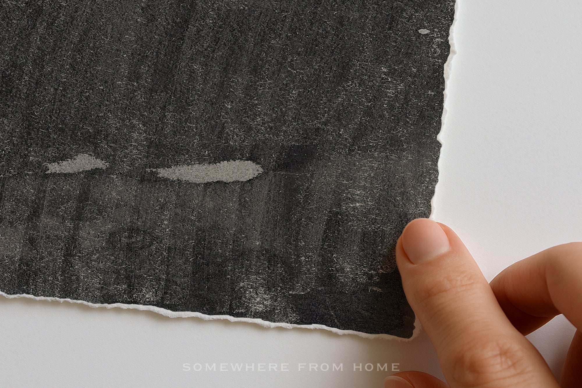 A close-up of a dark charcoal gray textured sheet of handmade paper with a rough, torn edge, held by a person's fingers against a clean white background.
