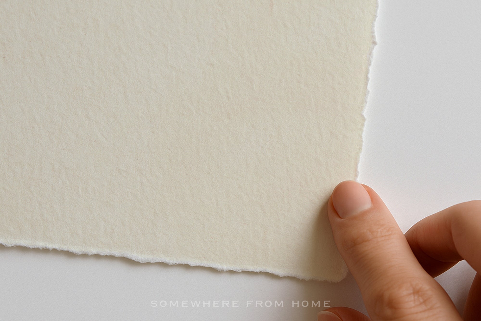 A close-up of a hand holding the edge of a textured off-white sheet of paper against a plain white background.