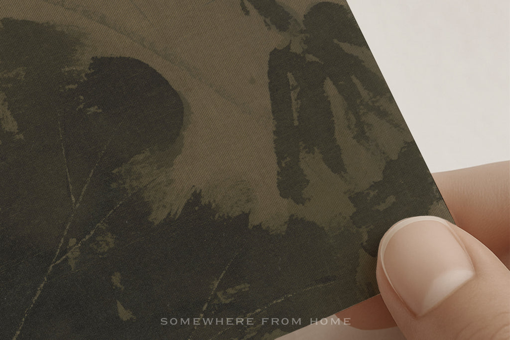 A close-up of a printed card or art print being held by fingers. The print features a dark, textured illustration in muted olive and black tones, with the text 'SOMEWHERE FROM HOME' near the bottom.