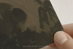 A close-up of a printed card or art print being held by fingers. The print features a dark, textured illustration in muted olive and black tones, with the text 'SOMEWHERE FROM HOME' near the bottom.