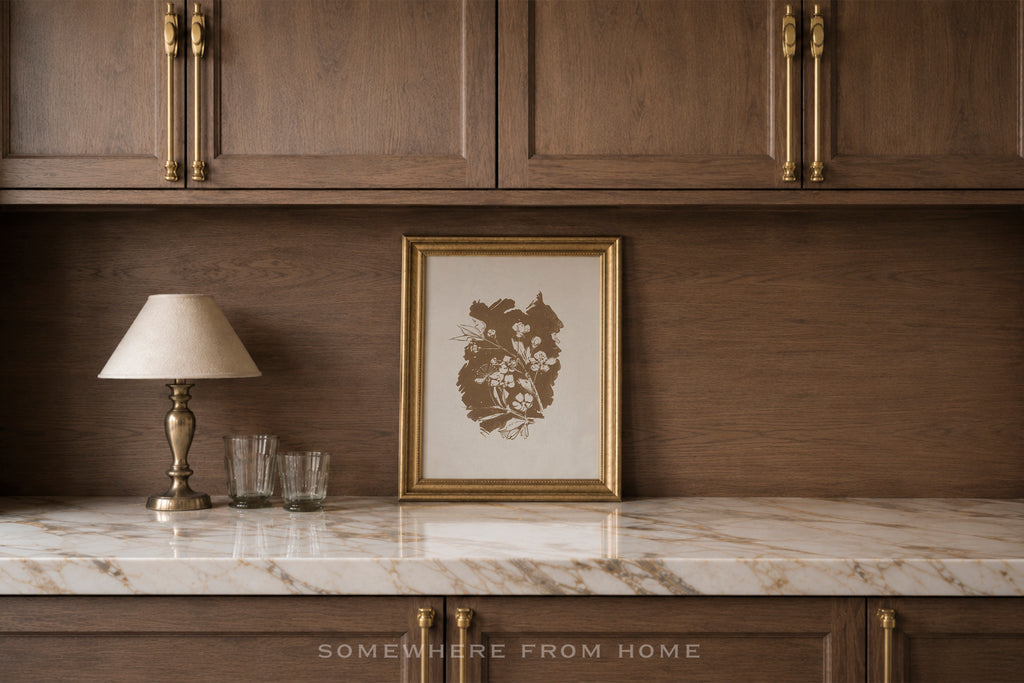 A framed botanical art print with a gold frame sits on a white marble countertop against a dark wood backsplash, next to a classic table lamp and two clear drinking glasses.