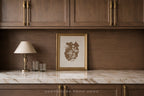 A framed botanical art print with a gold frame sits on a white marble countertop against a dark wood backsplash, next to a classic table lamp and two clear drinking glasses.