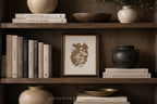 A framed botanical art print with a dark brown floral illustration on a cream background, styled on a dark wood shelf alongside books, ceramic vases, and bowls.