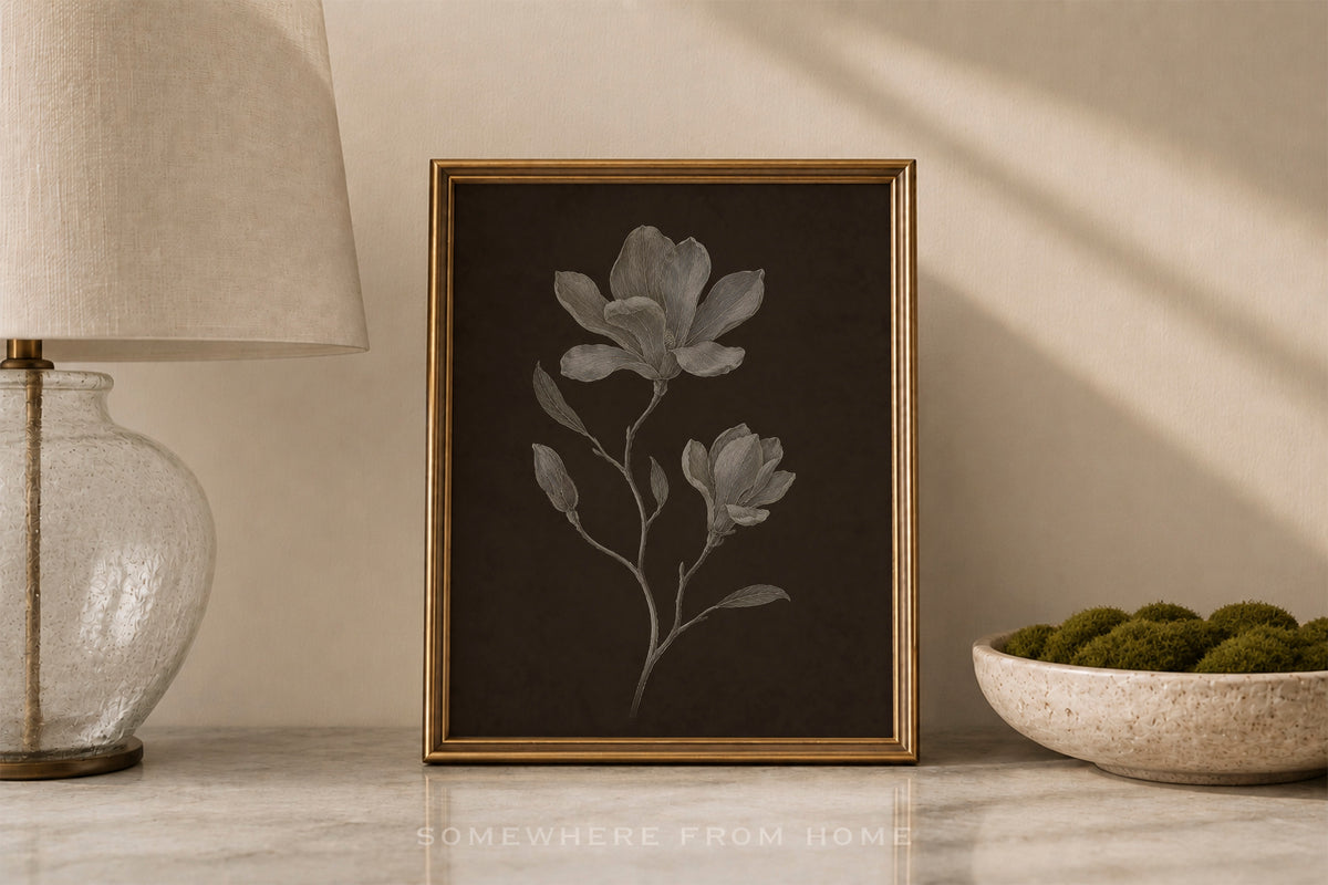 A framed botanical art print featuring light gray magnolia flowers on a dark brown background, styled on a marble surface next to a lamp and a bowl of moss.