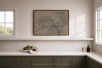 A framed botanical art print hangs on a light beige wall above a white shelf in a modern kitchen setting. The print features delicate, sketch-like tree branches in muted brown and gray tones.
