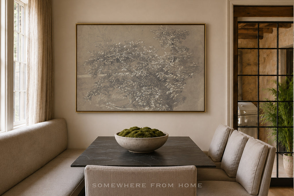 A large framed botanical art print with a gold frame hangs on a beige wall above a dining set in a softly lit room. The print depicts delicate tree branches with small white blossoms in muted gray and beige tones.
