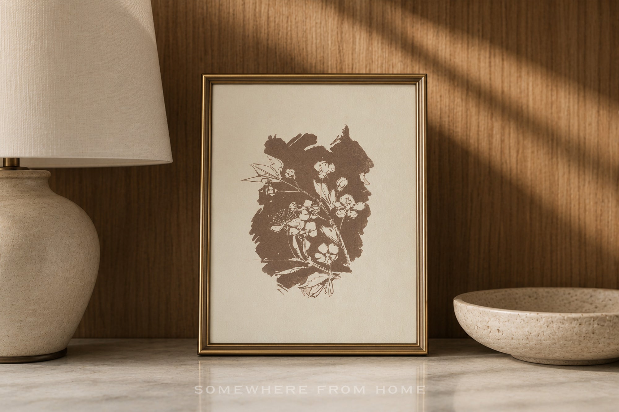 A framed botanical art print with a brown floral illustration on a cream background, set in a warm, styled interior next to a lamp and a ceramic bowl.