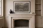 A framed black and white landscape art print hangs on a beige wall above a stone fireplace mantel, styled with a dark vase, a small figurine, and a white bowl. Built-in shelves are visible on the right.