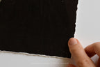 A close-up of a hand holding the edge of a large black sheet of torn paper against a white background, with the text 'SOMEWHERE FROM HOME' faintly visible at the bottom.
