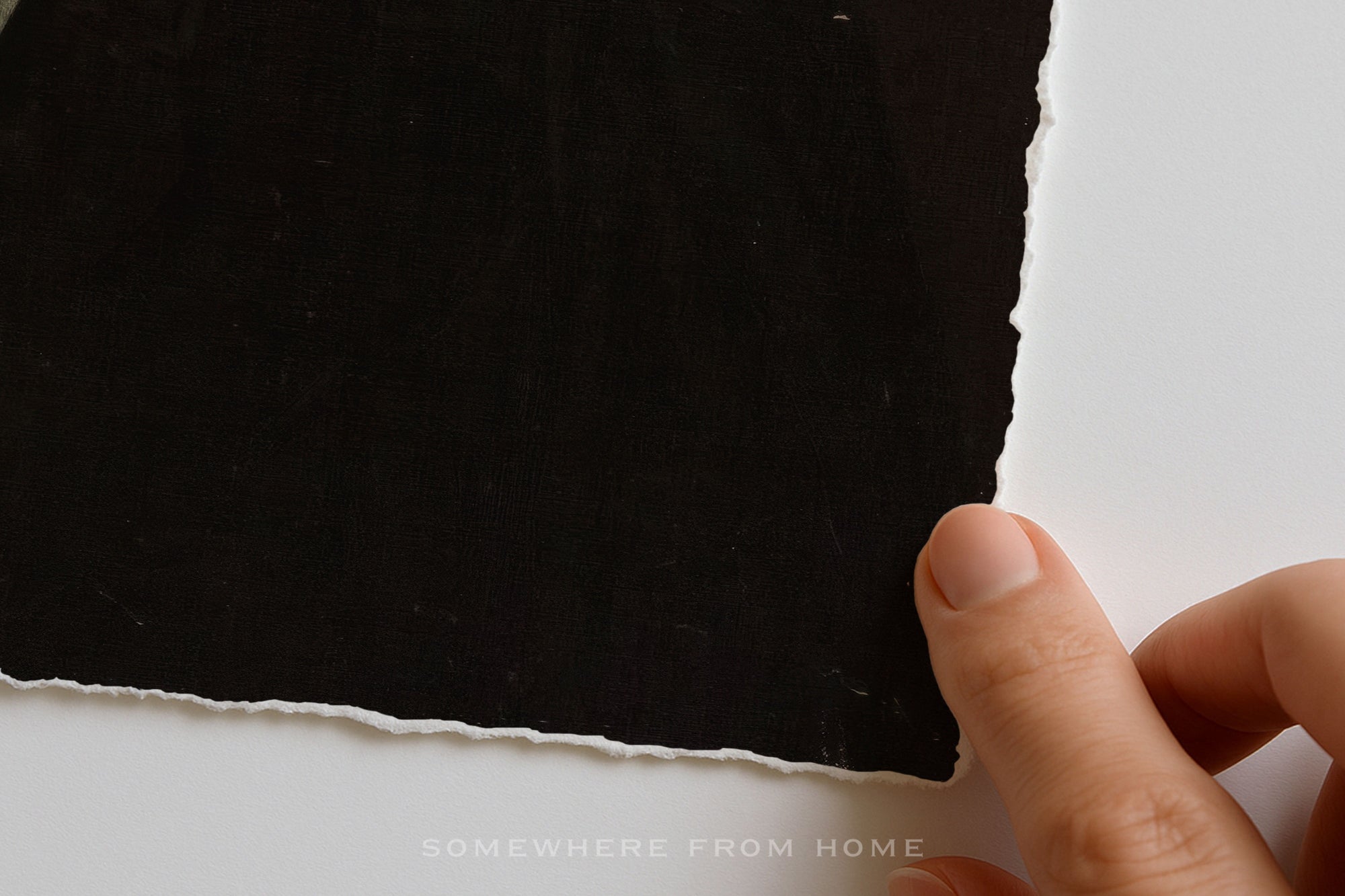 A close-up of a hand holding the edge of a large black sheet of torn paper against a white background, with the text 'SOMEWHERE FROM HOME' faintly visible at the bottom.