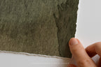 A close-up of a hand holding the edge of a square piece of textured, watercolor-like paper in a muted olive green shade against a plain white background.