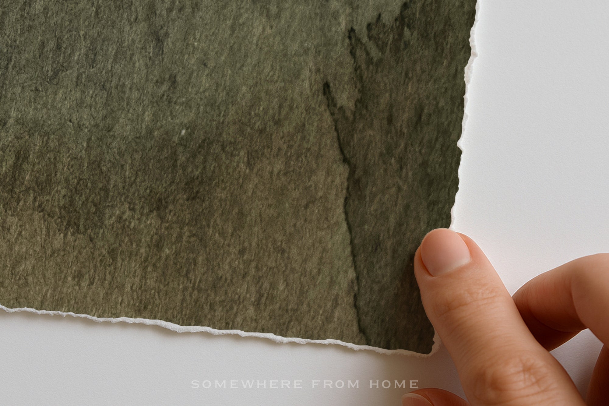 A close-up of a hand holding the edge of a square piece of textured, watercolor-like paper in a muted olive green shade against a plain white background.