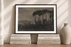 A framed sepia-toned landscape art print leaning against a wall on a wooden surface, styled with stacked books and a ceramic vase in a warm interior setting.