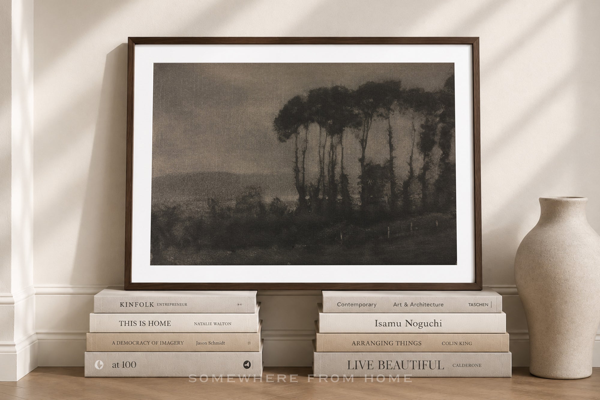 A framed sepia-toned landscape art print leaning against a wall on a wooden surface, styled with stacked books and a ceramic vase in a warm interior setting.