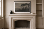A framed black and white landscape art print hangs on a beige wall above a stone fireplace mantel, styled with a dark vase and a small white bowl in a classic interior setting.