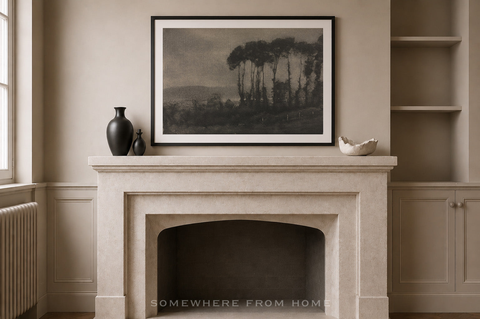 A framed black and white landscape art print hangs on a beige wall above a stone fireplace mantel, styled with a dark vase and a small white bowl in a classic interior setting.
