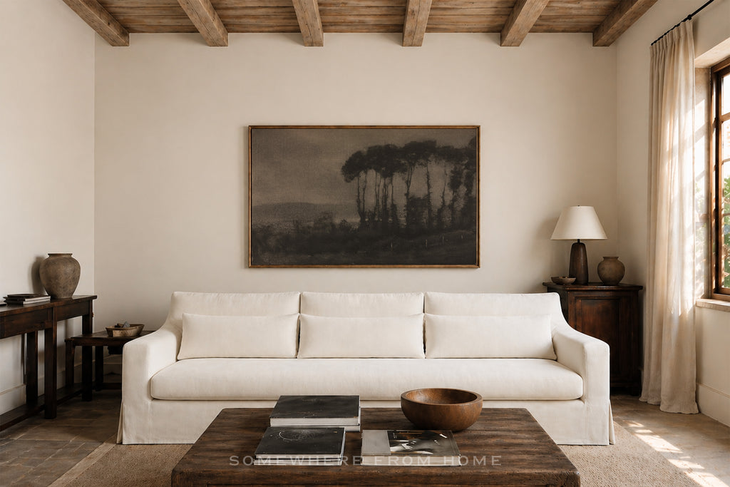 A large framed landscape art print depicting pine trees in a misty forest, displayed on a cream wall above a white fabric sofa in a rustic, sunlit living room.