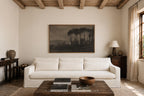 A large framed landscape art print depicting pine trees in a misty forest, displayed on a cream wall above a white fabric sofa in a rustic, sunlit living room.