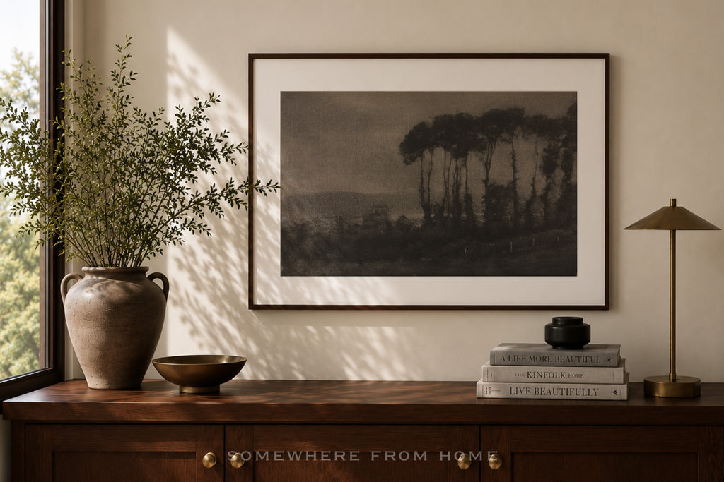A framed landscape art print hangs on a beige wall above a dark wood console table. The print depicts a moody, dark-toned scene of tall trees and a distant valley.
