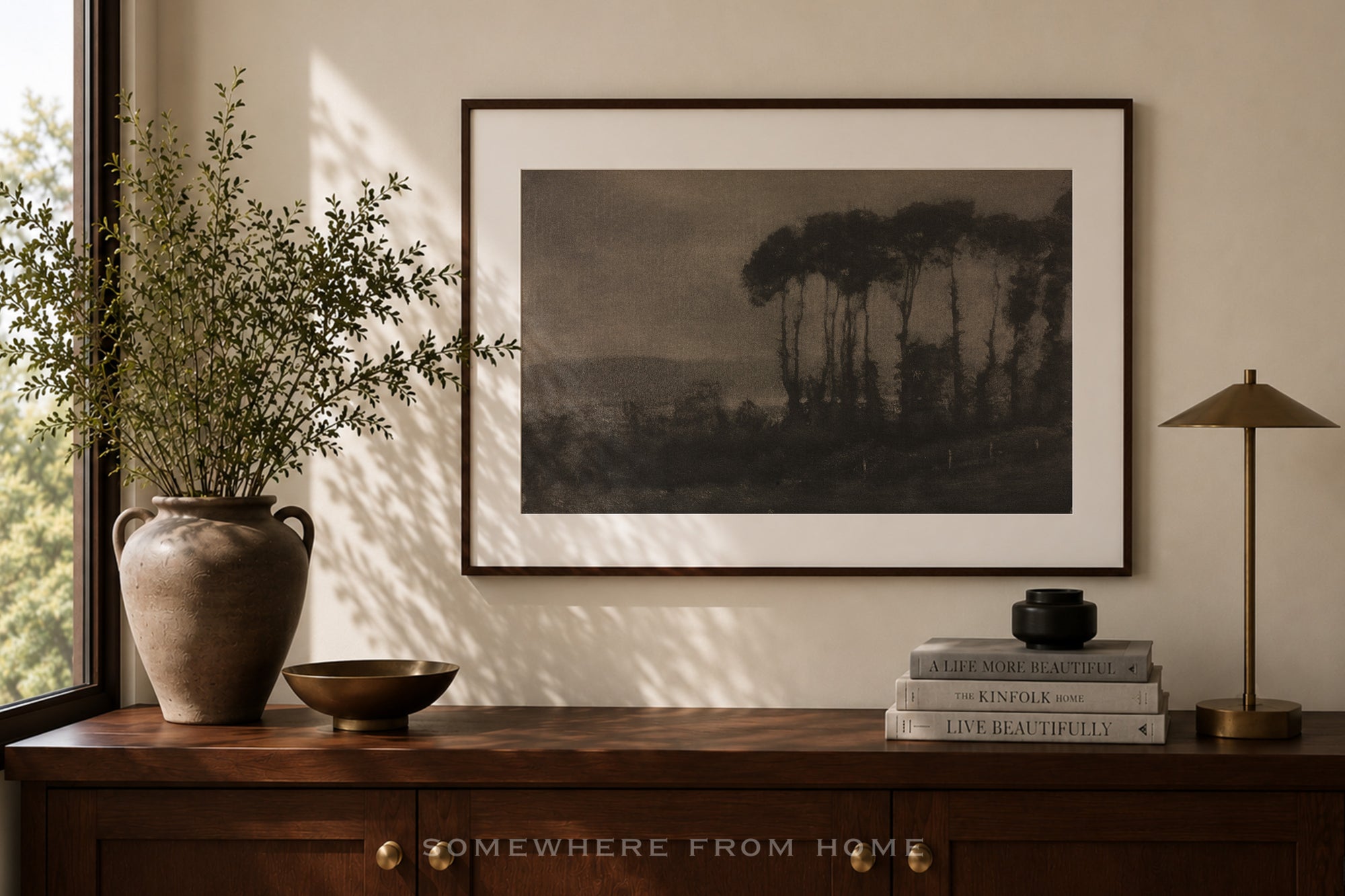 A framed landscape art print hangs on a beige wall above a dark wood console table. The print depicts a moody, dark-toned scene of tall trees and a distant valley.