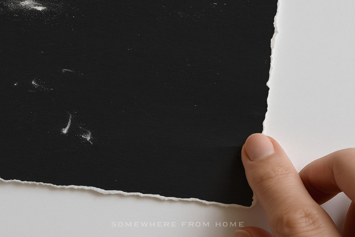 A close-up of a hand holding the edge of a black textured sheet of torn paper against a white background. The bottom of the image features a faint watermark reading 'SOMEWHERE FROM HOME'.