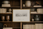 A framed black and white landscape sketch with elegant cursive writing, set in a gold frame and displayed on a dark wood bookshelf surrounded by books and decorative pottery.