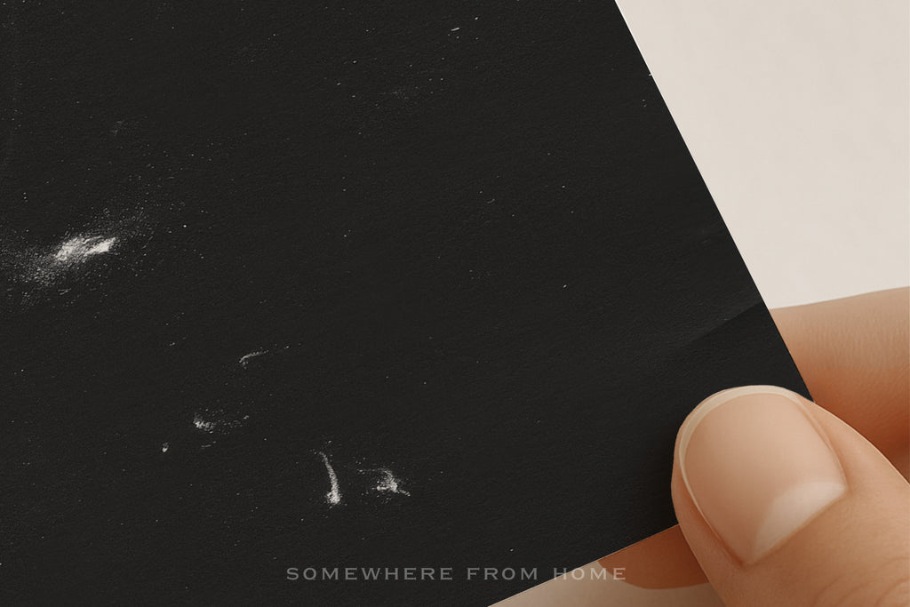 A close-up of a hand holding a black rectangular item, likely a phone case or card, against a plain light background. The black surface has a few small white scratch-like marks and a faint 'SOMEWHERE FROM HOME' watermark near the bottom.