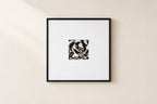 A square black-framed wall art print with a wide white mat, featuring a small central black-and-white abstract pattern, hung on a warm beige wall.
