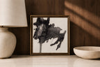 A framed abstract black ink painting on textured off-white paper, styled on a marble surface between a ceramic lamp and a shallow bowl against a dark wood wall.