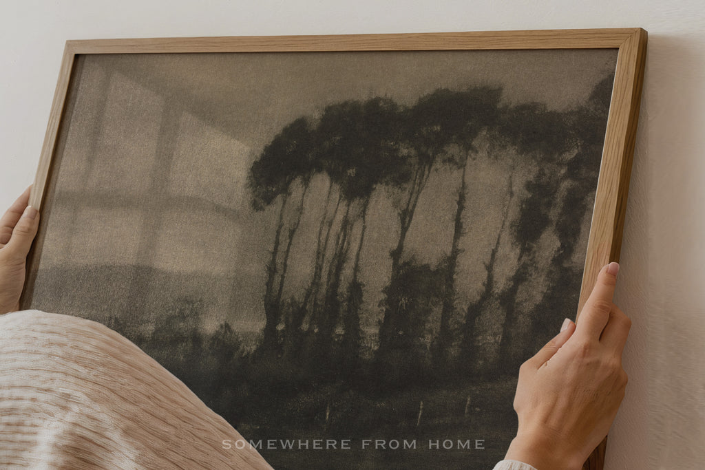 A person holds a framed art print featuring a dark, moody forest scene with tall trees. The print has a textured, sepia-toned look and is housed in a light natural wood frame.