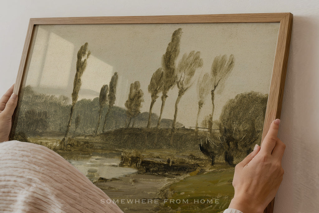 A framed landscape art print being held by two hands against a plain wall. The artwork depicts a muted, sepia-toned scene with tall trees, hills, and a river, enclosed in a light wood frame.