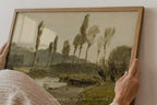 A framed landscape art print being held by two hands against a plain wall. The artwork depicts a muted, sepia-toned scene with tall trees, hills, and a river, enclosed in a light wood frame.