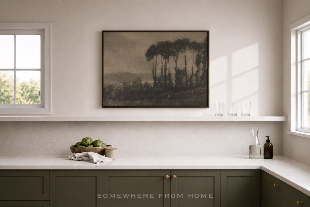 A framed landscape art print featuring dark trees and hills, displayed on a white wall above a kitchen counter in a modern, neutral-toned room.