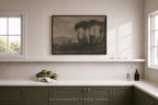 A framed landscape art print featuring dark trees and hills, displayed on a white wall above a kitchen counter in a modern, neutral-toned room.