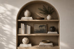 An arched wooden built-in bookshelf with three shelves, styled with ceramic vases, books, a small bowl, and a framed landscape print, set against a warm beige wall with soft leaf shadows.
