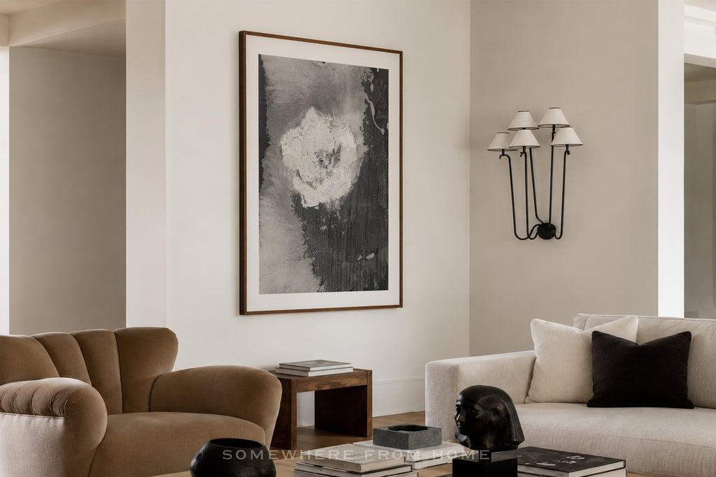 A framed abstract black and white wall art print hangs on a beige wall in a modern living room setting with a brown armchair, a white sofa, and a black wall sconce.