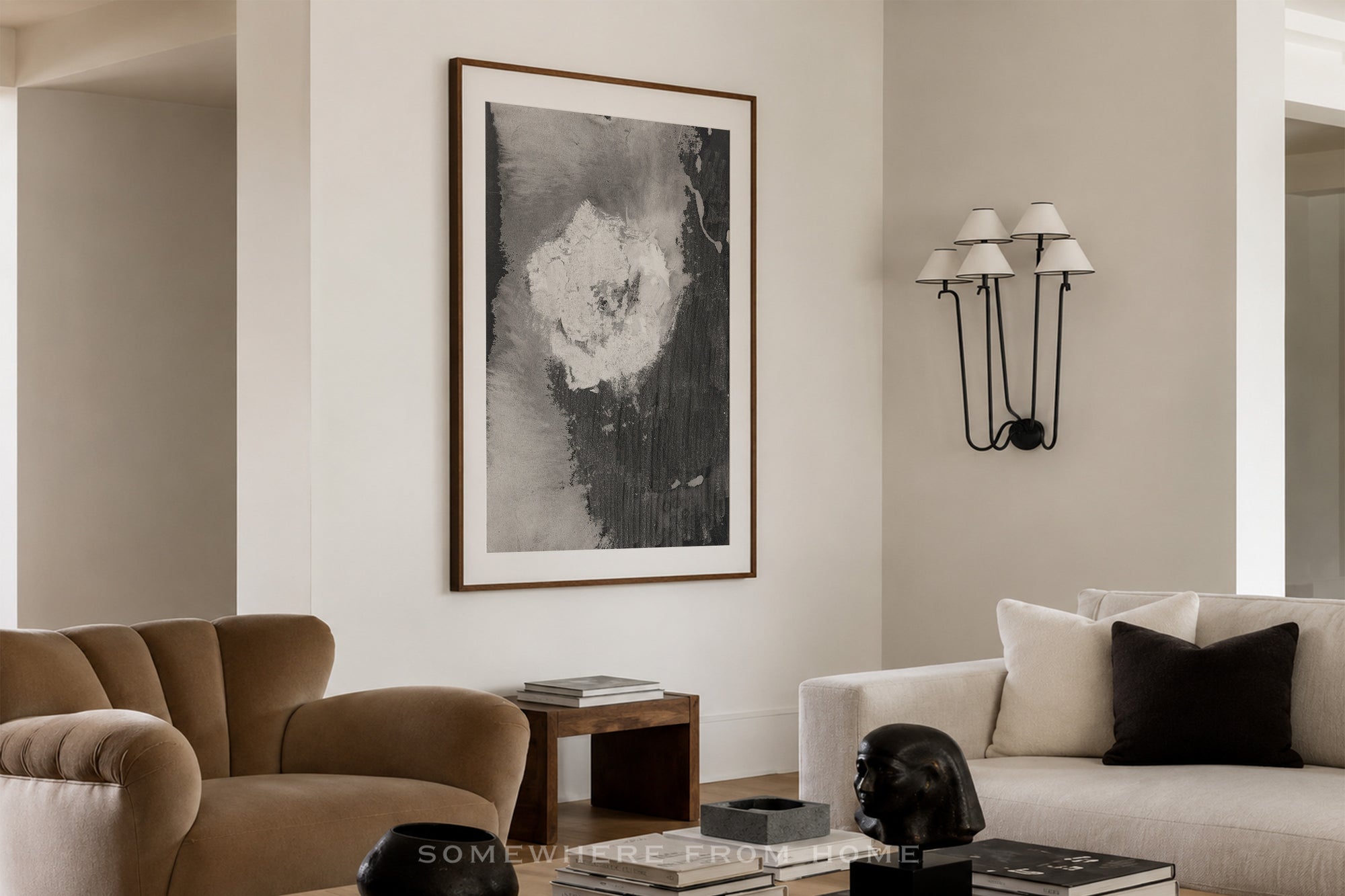 A framed abstract black and white wall art print hangs on a beige wall in a modern living room setting with a brown armchair, a white sofa, and a black wall sconce.