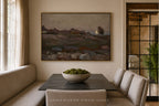 A framed landscape painting hangs on a beige wall above a dark rectangular table in a dining area with upholstered bench seating and a bowl of green moss on top. The scene is styled as a warm, modern interior.