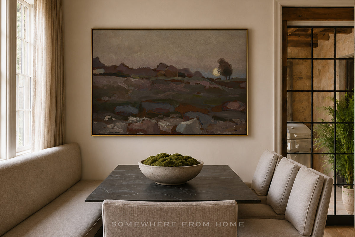 A framed landscape painting hangs on a beige wall above a dark rectangular table in a dining area with upholstered bench seating and a bowl of green moss on top. The scene is styled as a warm, modern interior.
