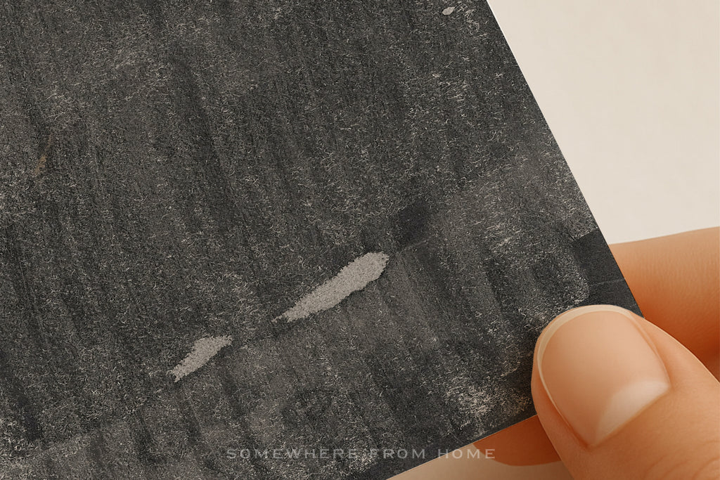 A close-up of a hand holding a textured charcoal or black slate card with a white background visible in the corner. The card has a rough, brushed texture with two small white scratch-like marks.