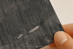 A close-up of a hand holding a textured charcoal or black slate card with a white background visible in the corner. The card has a rough, brushed texture with two small white scratch-like marks.