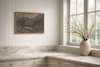 A framed landscape painting hangs on a white paneled wall above a marble countertop in a bright kitchen. A textured ceramic vase with delicate branches sits on the windowsill.