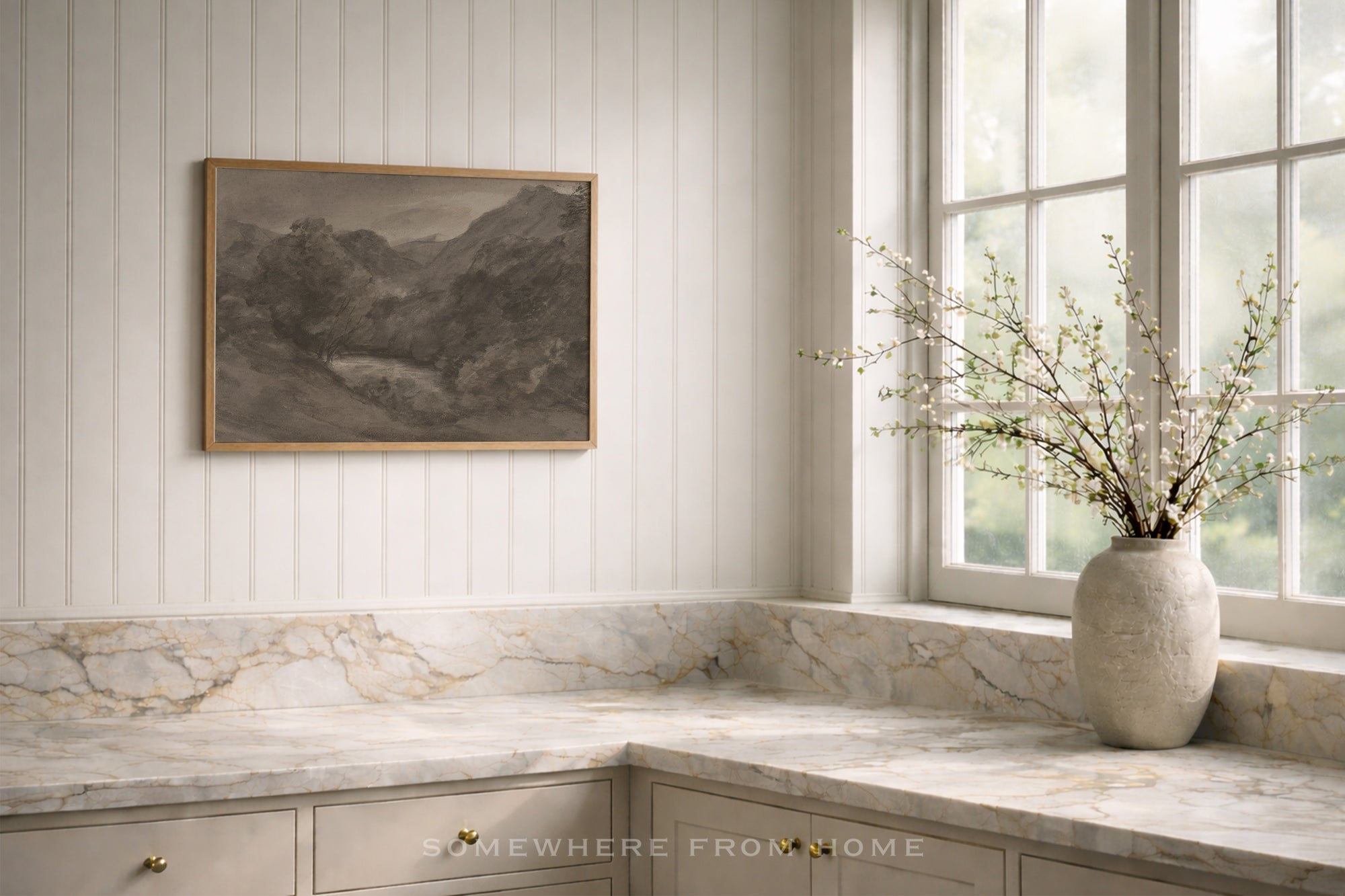 A framed landscape painting hangs on a white paneled wall above a marble countertop in a bright kitchen. A textured ceramic vase with delicate branches sits on the windowsill.