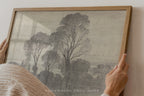 A framed art print featuring a monochrome sketch of trees, held by hands against a light-colored wall. The print is enclosed in a light wood frame.