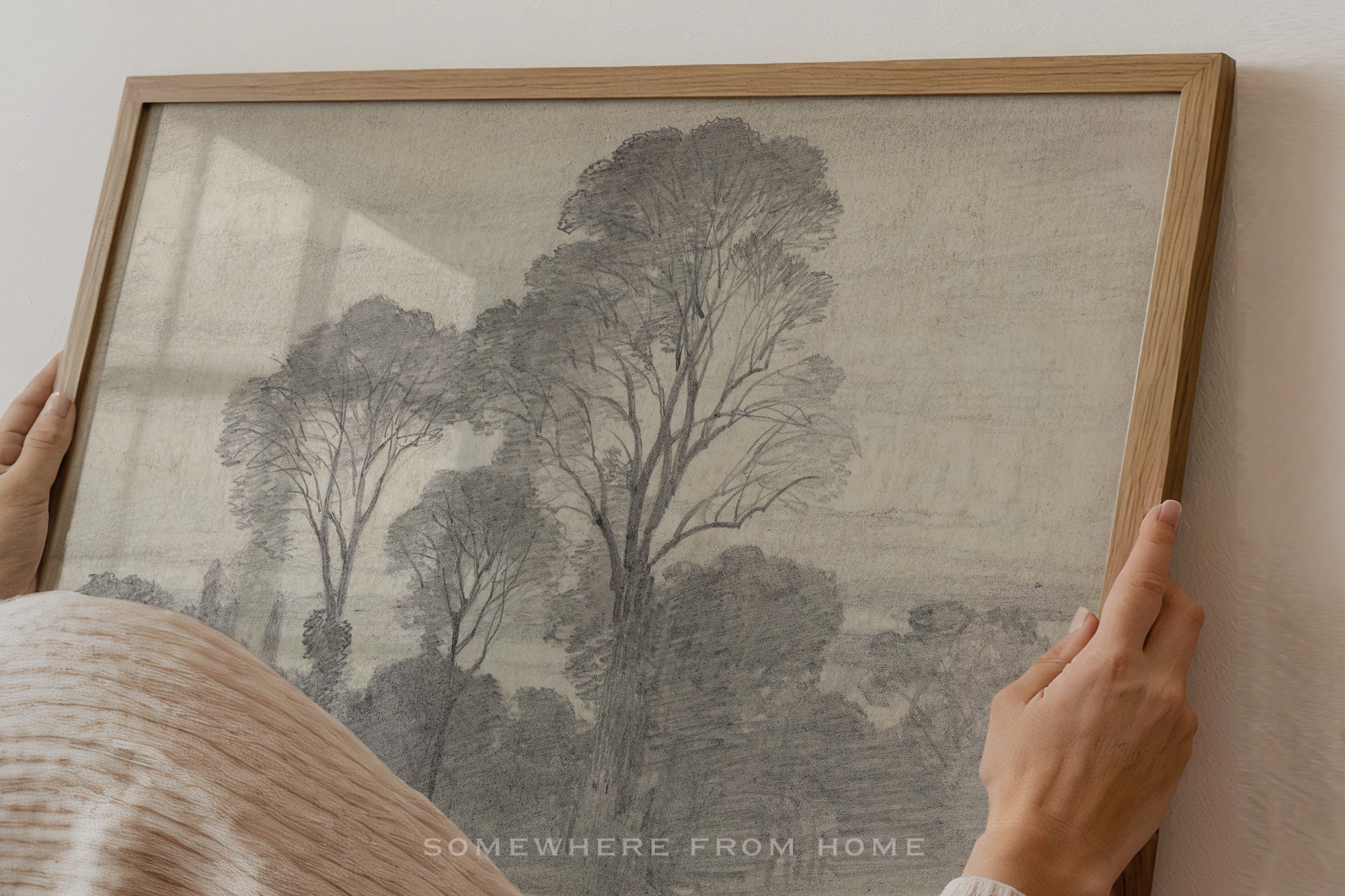 A framed art print featuring a monochrome sketch of trees, held by hands against a light-colored wall. The print is enclosed in a light wood frame.