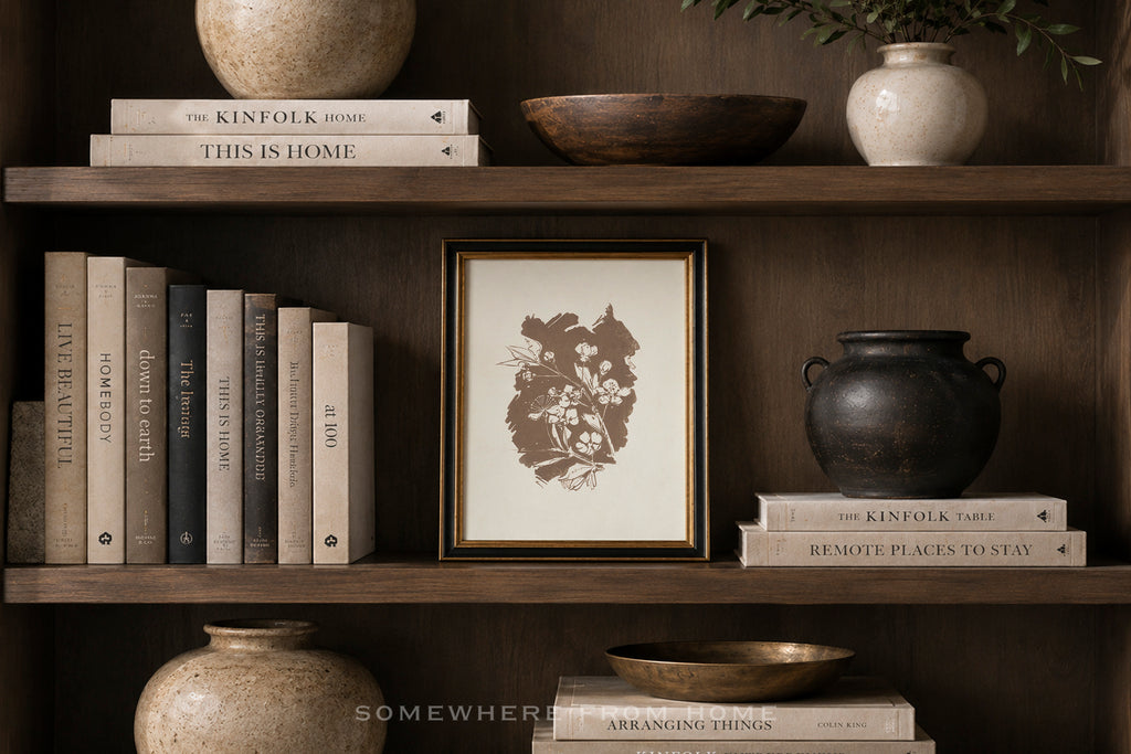 A framed botanical art print with a dark brown floral illustration on a cream background, styled on a dark wood shelf alongside books, ceramic vases, and bowls.