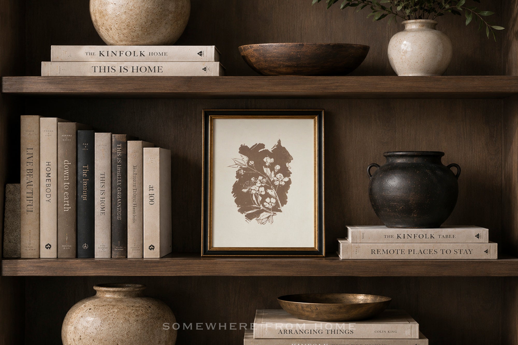 A framed botanical art print with a dark brown floral illustration on a cream background, styled on a dark wood shelf alongside books, ceramic vases, and bowls.